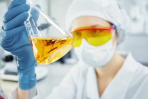 Close up portrait of young woman's hands holding test tube with yellow liquid. Medical researcher or doctor wearing surgical mask and safety glasses looking at glass tube while working in laboratory